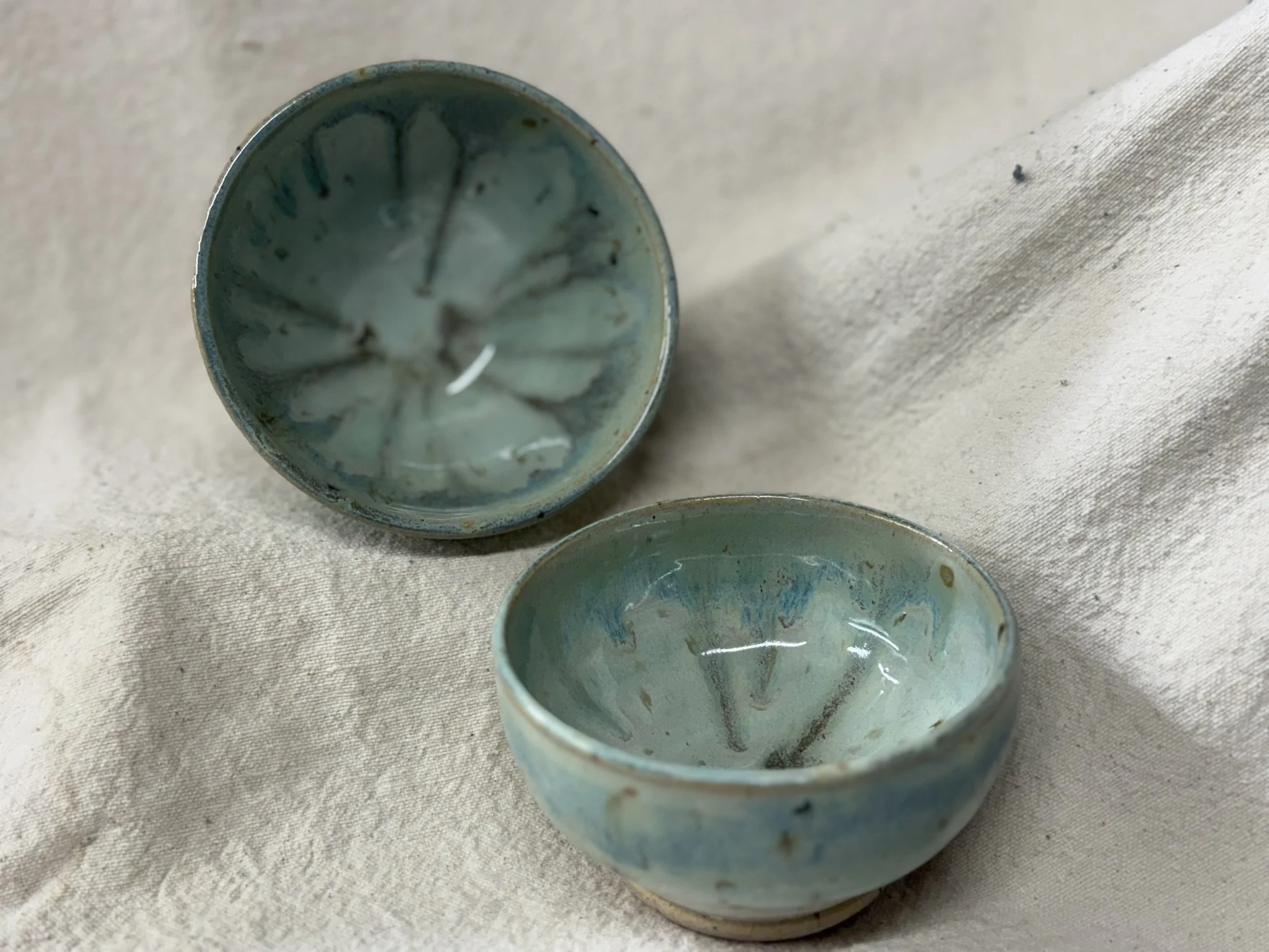 Two small ceramic bowls with a turquoise glaze, one standing upright and the other on its side, on a beige fabric surface.