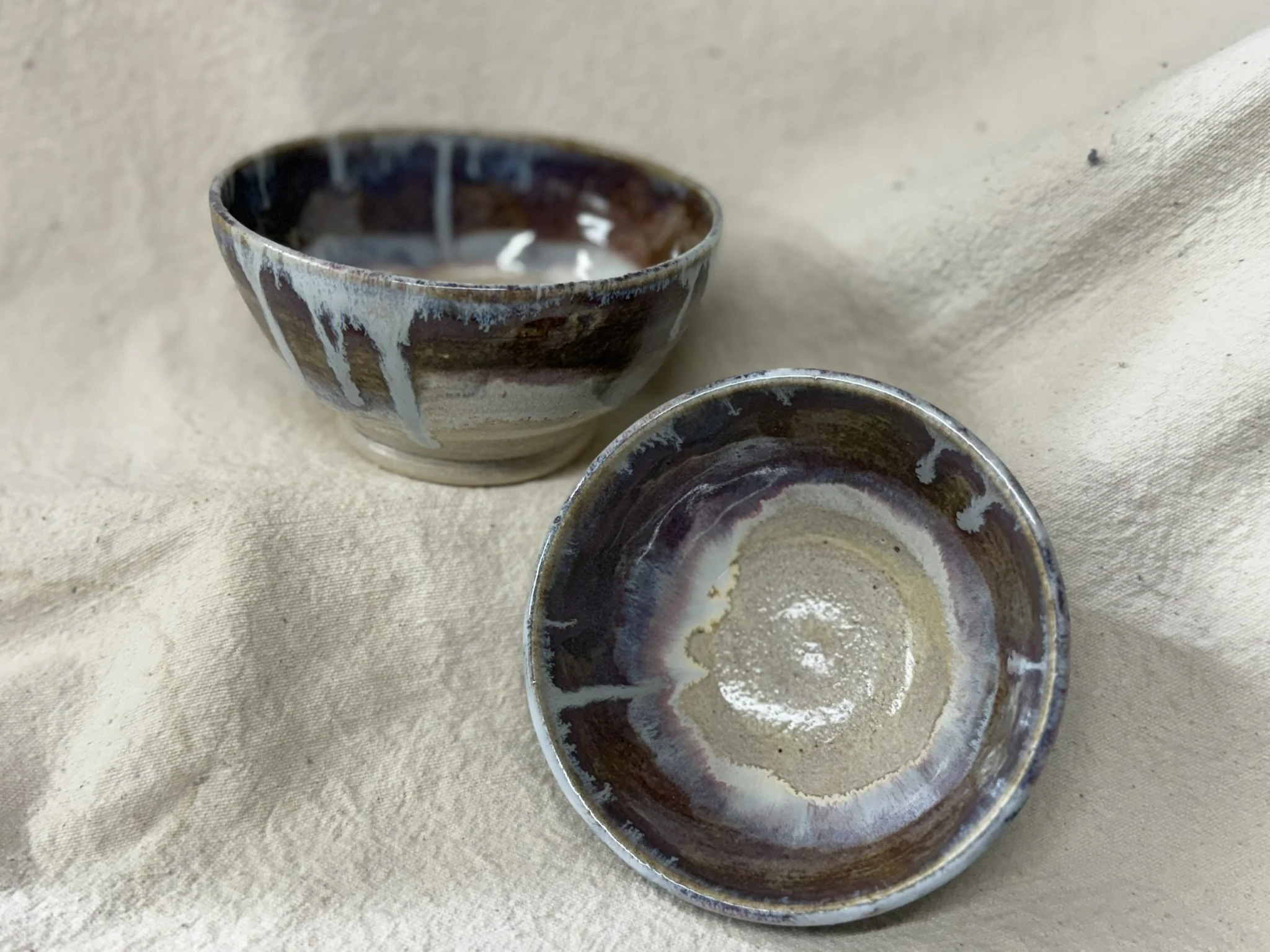 Two small ceramic bowls with brown, cream, and blue glaze, placed on a beige fabric surface, one bowl with a creamy substance inside.