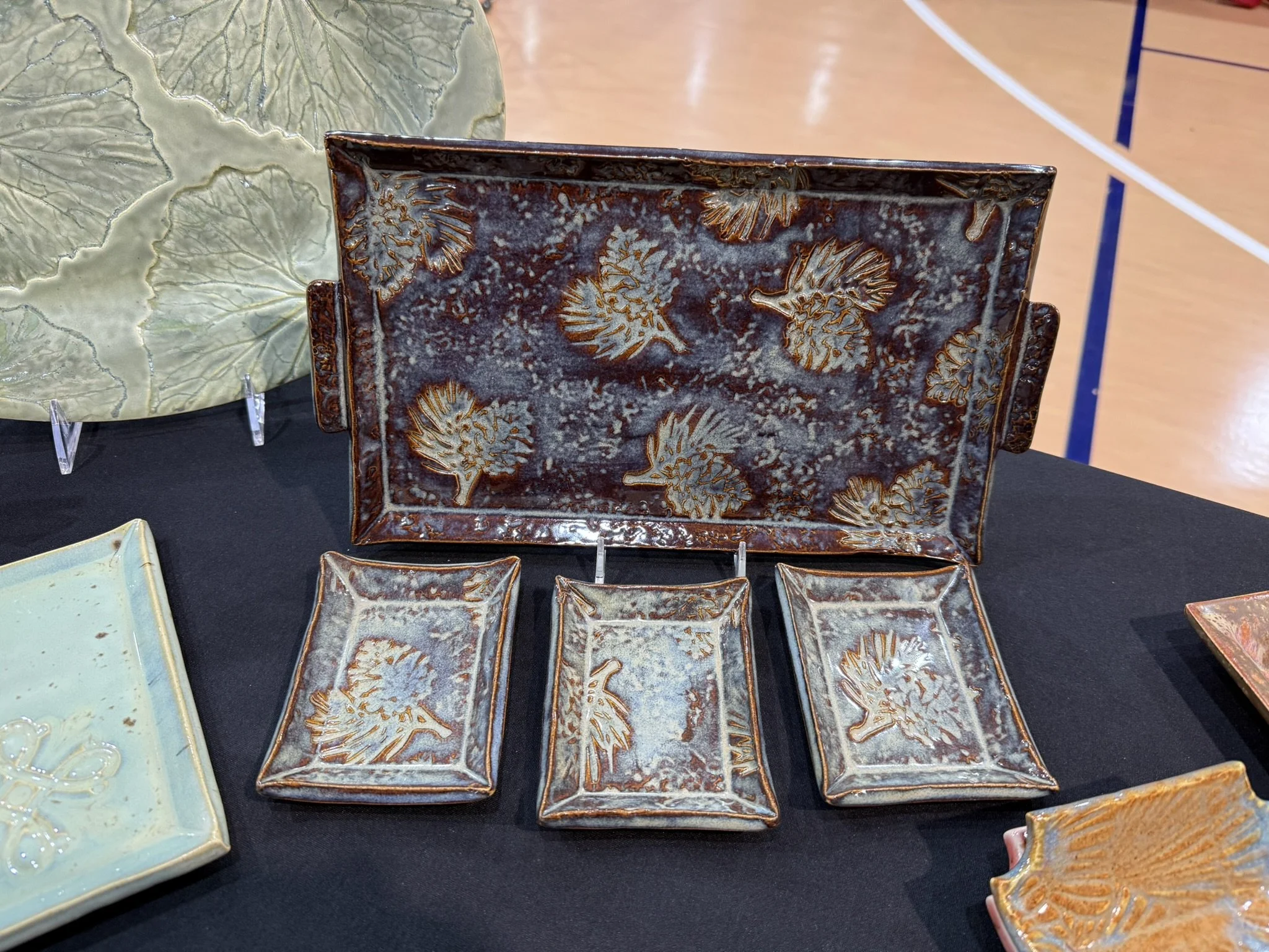 Set of handcrafted ceramic plates with leaf patterns, displayed on a black table, with a sports court in the background.