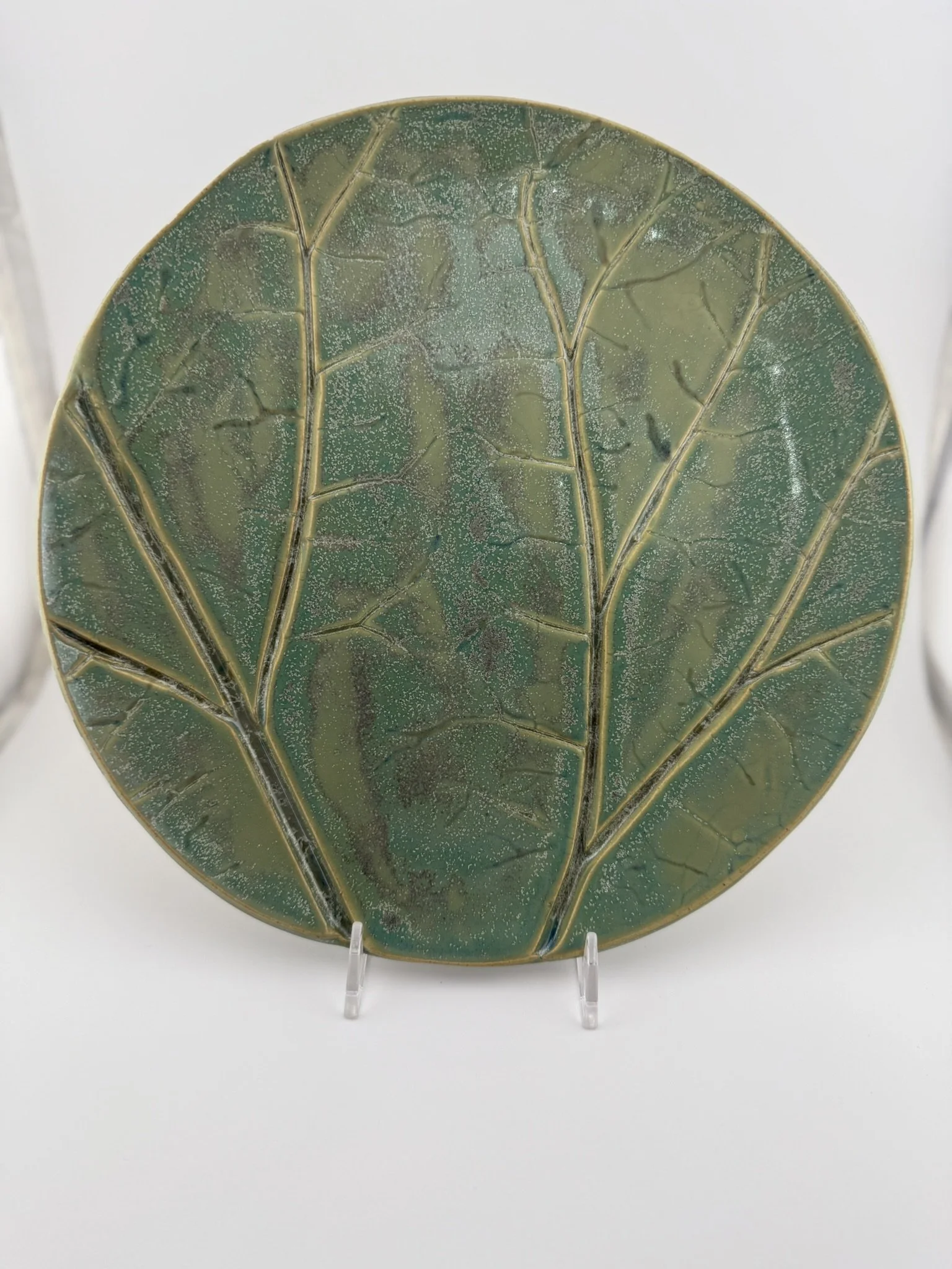 Decorative ceramic plate with a green and brown leaf pattern, displayed on a clear stand.
