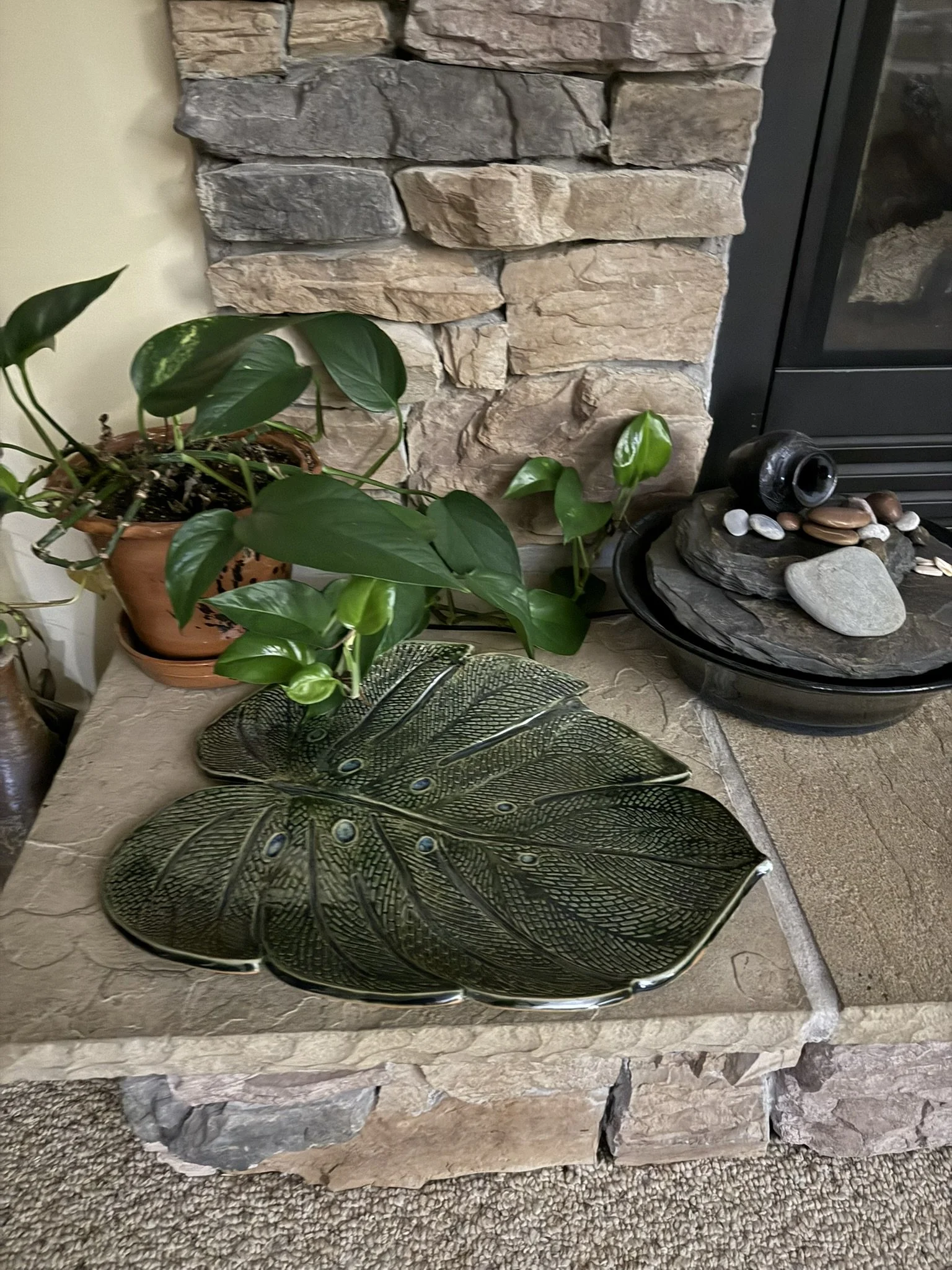 Decorative scene with potted green plants, a large leaf-shaped ceramic dish on a stone table, and a black bowl with rocks and a vase on top, set against a stone fireplace and black framed glass door.