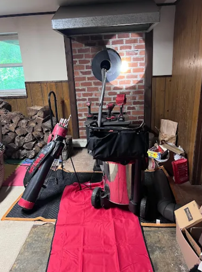 A telescope and a red chair inside a room with wood panel walls and a brick fireplace, with firewood stacked on the left side and a pile of cardboard boxes on the right.