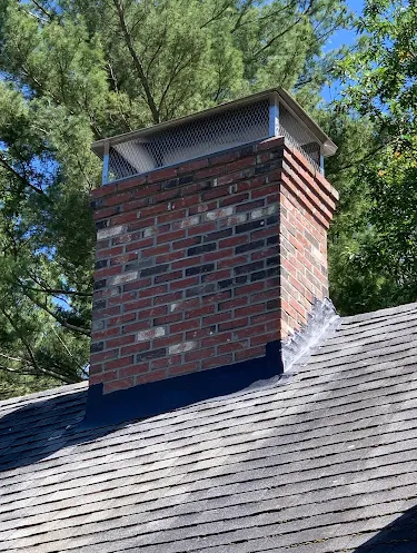 A brick chimney on a house's roof with a metal cap, surrounded by trees and a clear blue sky.