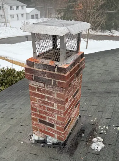 Brick chimney with a mesh cage and a concrete cap on a roof, with snow in the background.