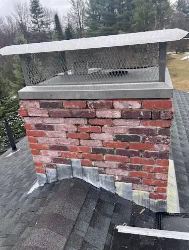 A brick chimney with a metal and mesh cap, sitting on a shingled roof with trees in the background.