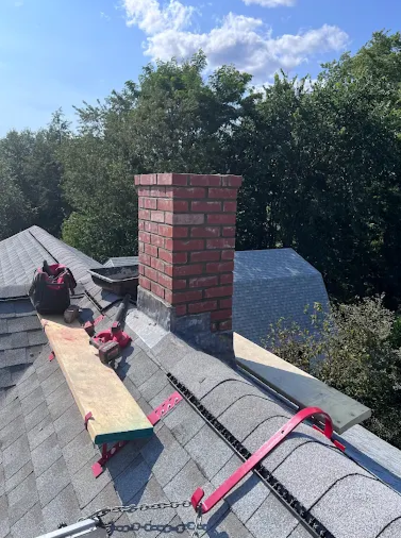 View of a roof with a brick chimney, roofing tools, and equipment, and surrounding trees under a partly cloudy sky.