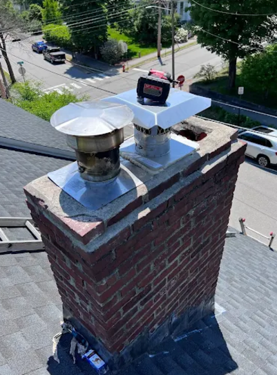 View of a brick chimney with two metal vent pipe caps, a metal chimney cap, and a level tool on top, as seen from a rooftop.
