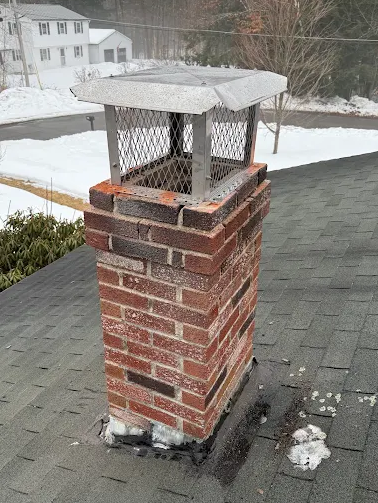 Brick chimney with metal cap on a shingles roof in winter, with snow on the ground and houses in the background.
