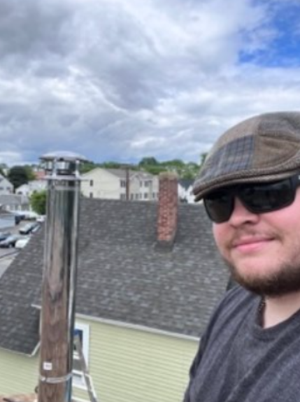 A man wearing a flat cap and sunglasses taking a selfie on a rooftop with a residential neighborhood and cloudy sky in the background.