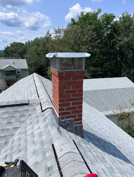 Close-up of a residential roof with a red brick chimney and a metal cap, surrounded by trees and neighboring houses.