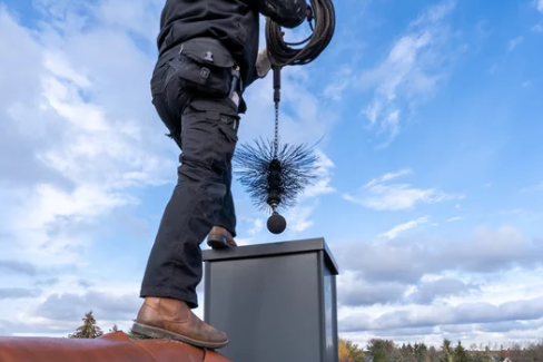 Person standing on a rooftop, installing or inspecting a black sphere and spiky object on a metal box, with a partly cloudy sky in the background.