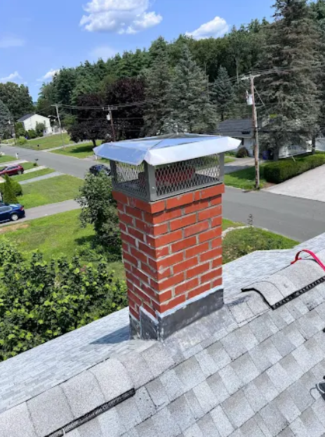 A chimney with a metal cap and a brick base on a residential house roof, with trees, houses, and a street in the background.