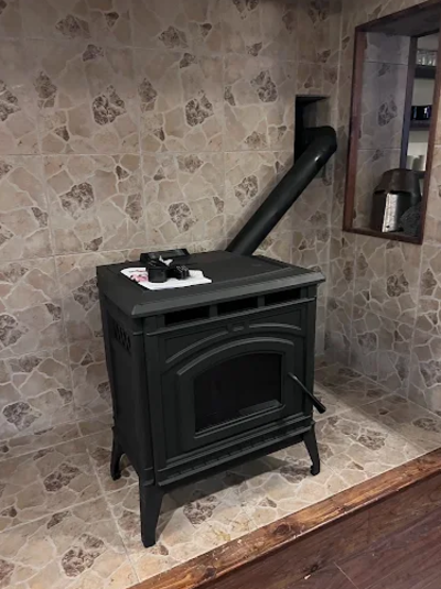 A black wood stove with a chimney pipe, positioned against a stone-tiled wall in a room with a wooden floor and a small window.