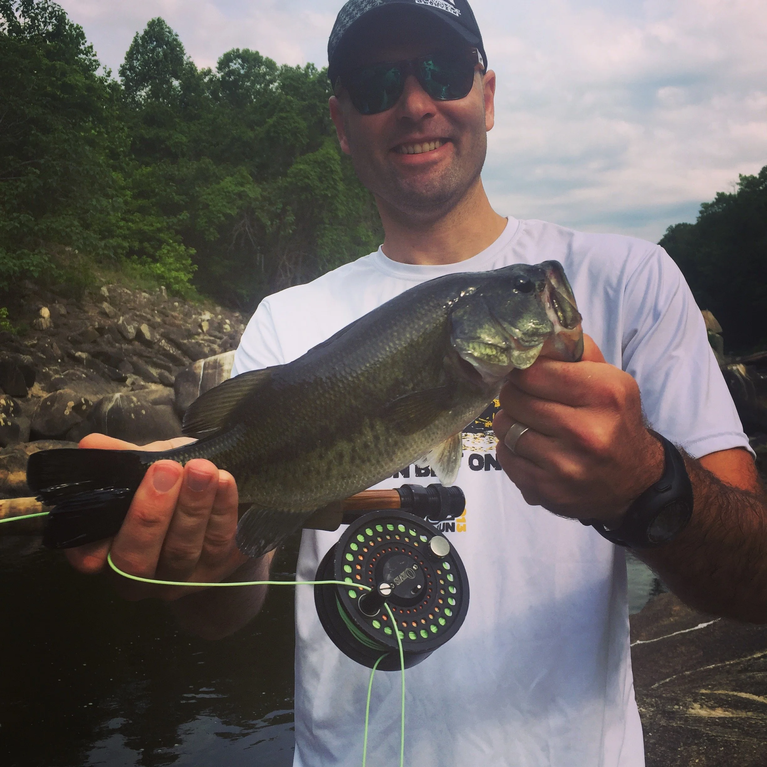 A man wearing sunglasses, a white t-shirt, and a black cap, holding a large fish with both hands near a rocky riverbank with trees in the background.