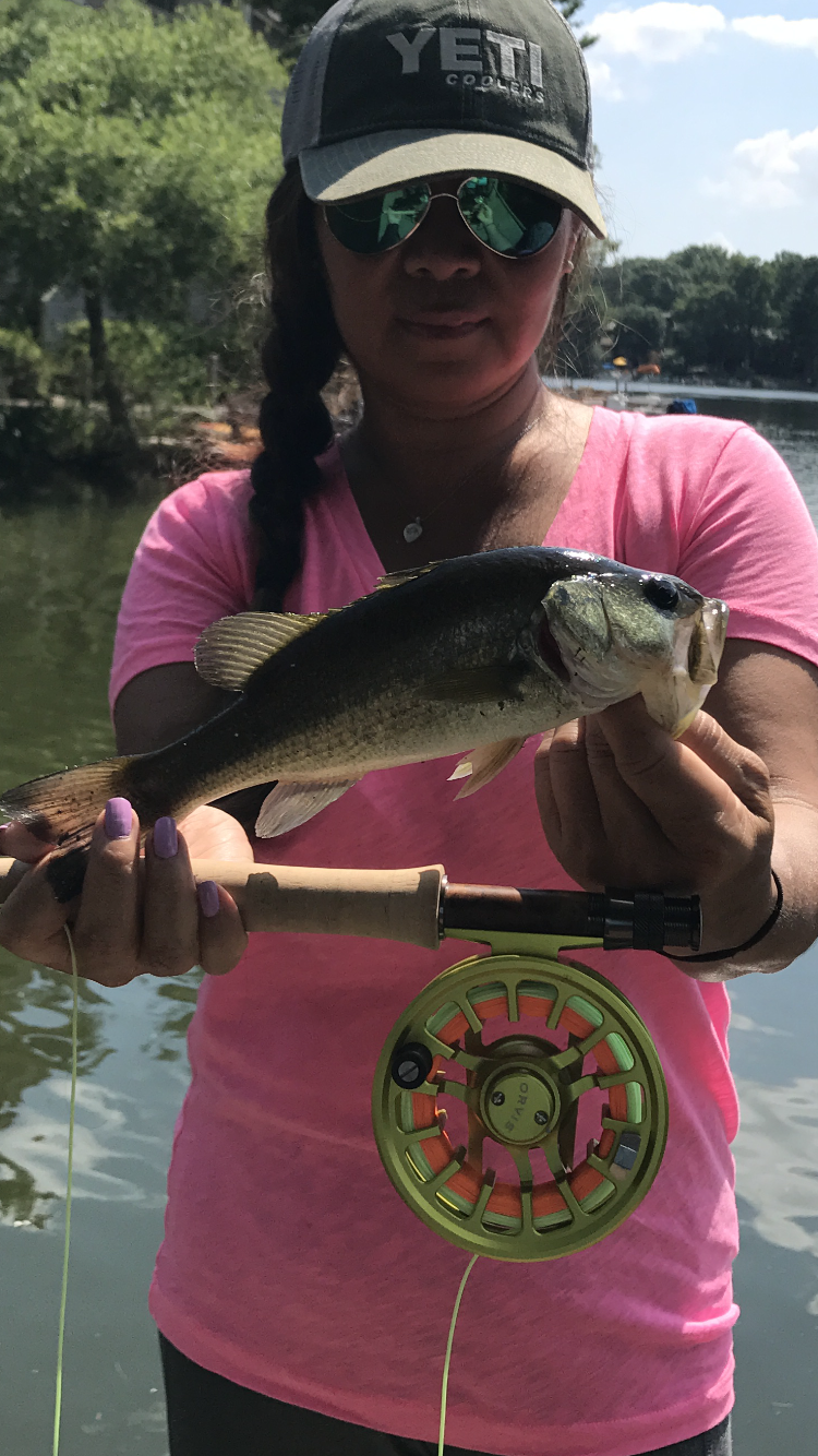 A woman wearing a pink shirt, sunglasses, and a cap holding a fishing rod with a fish she caught outdoors by a lake with trees in the background.