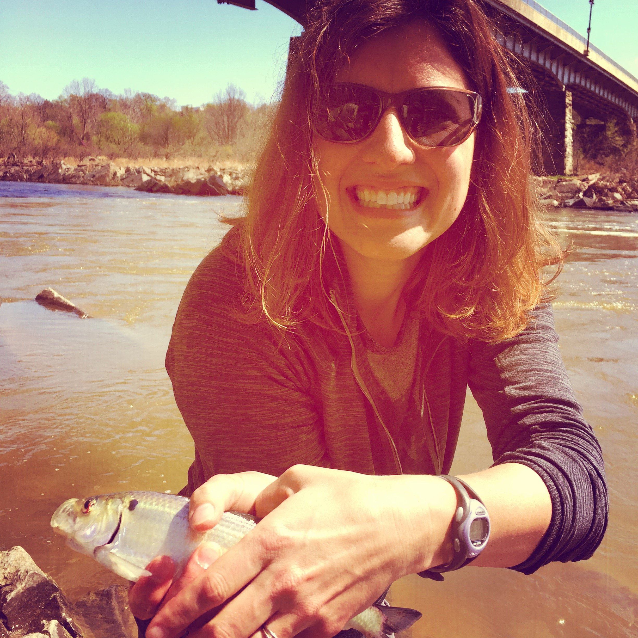 Woman with sunglasses smiling, holding a fish near a riverbank with a bridge in the background.