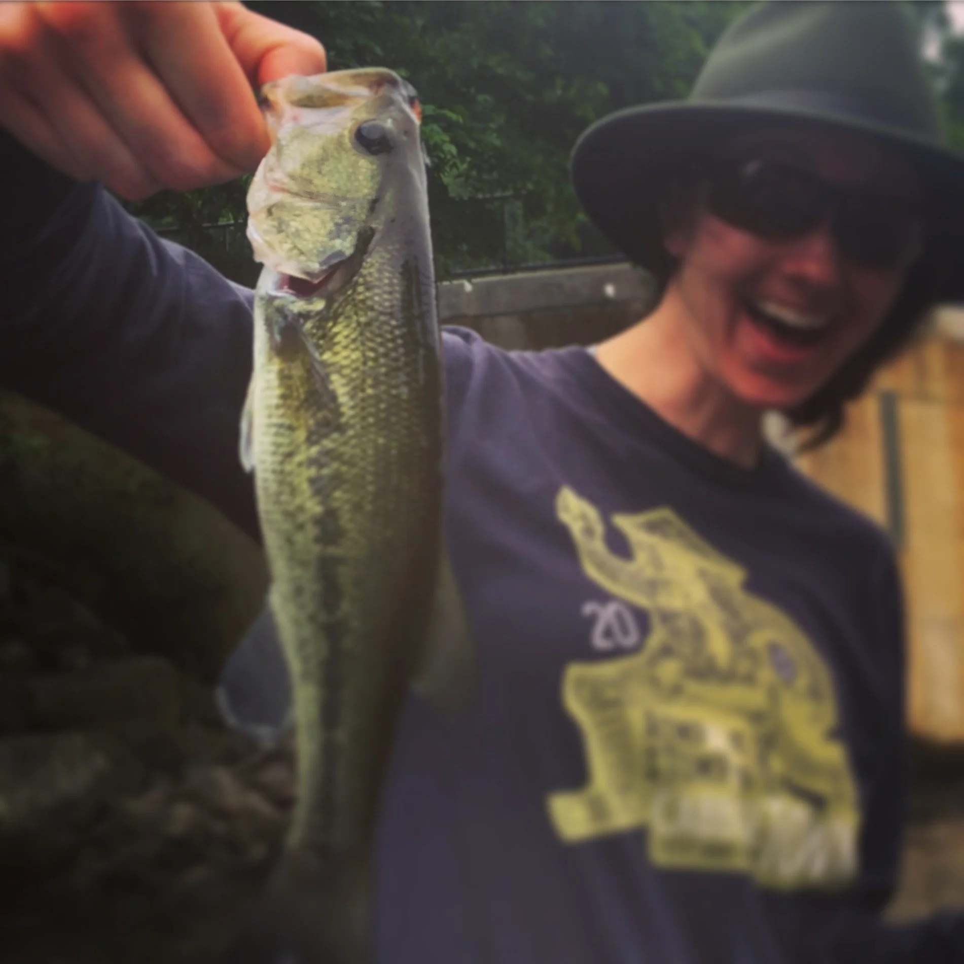 A woman wearing a black hat and sunglasses holding a largemouth bass with a smile.