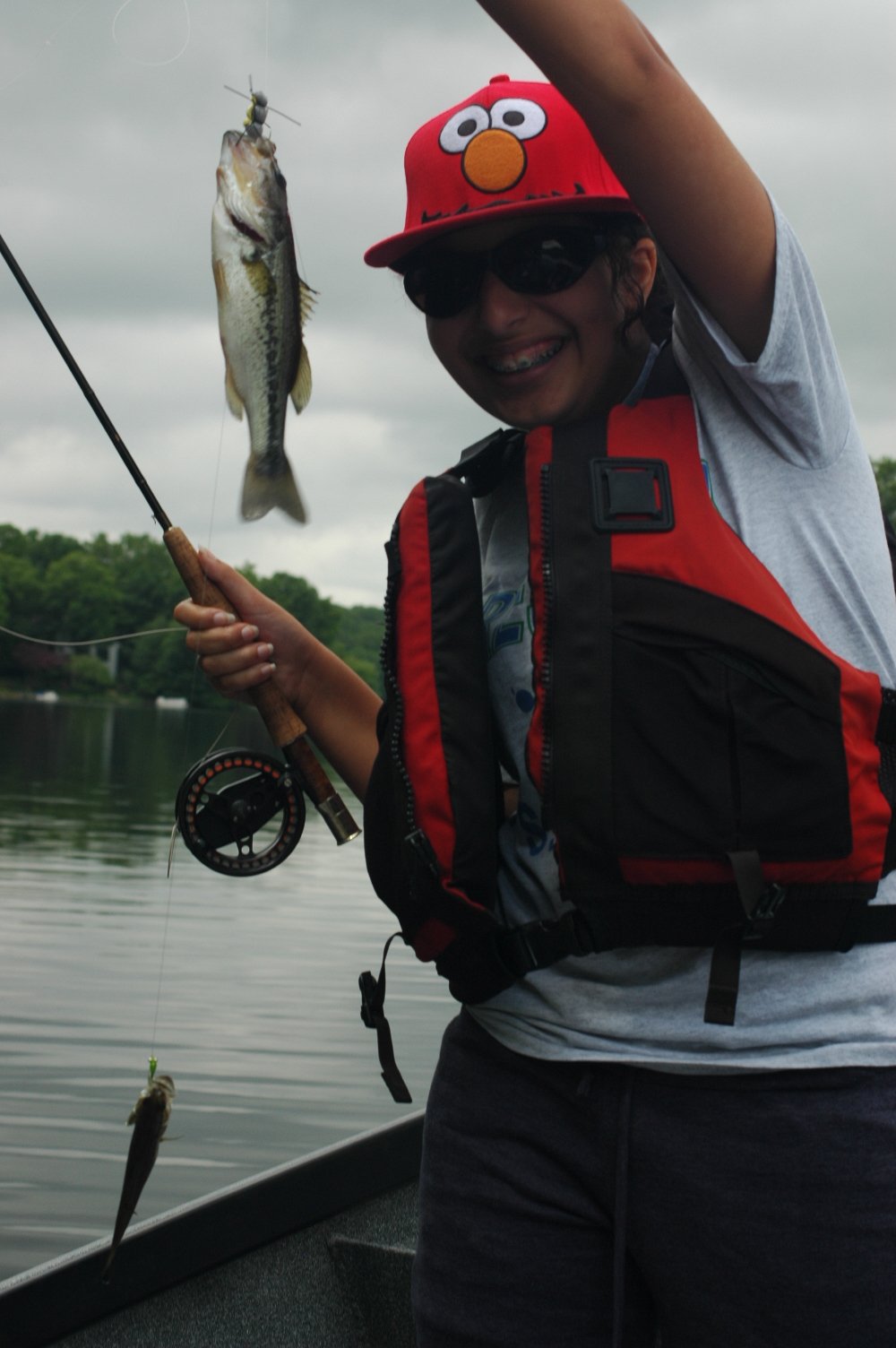A woman holding a fishing rod with a freshly caught fish, wearing sunglasses, a red life jacket, and a red hat with a cartoon face, on a lake with cloudy skies.