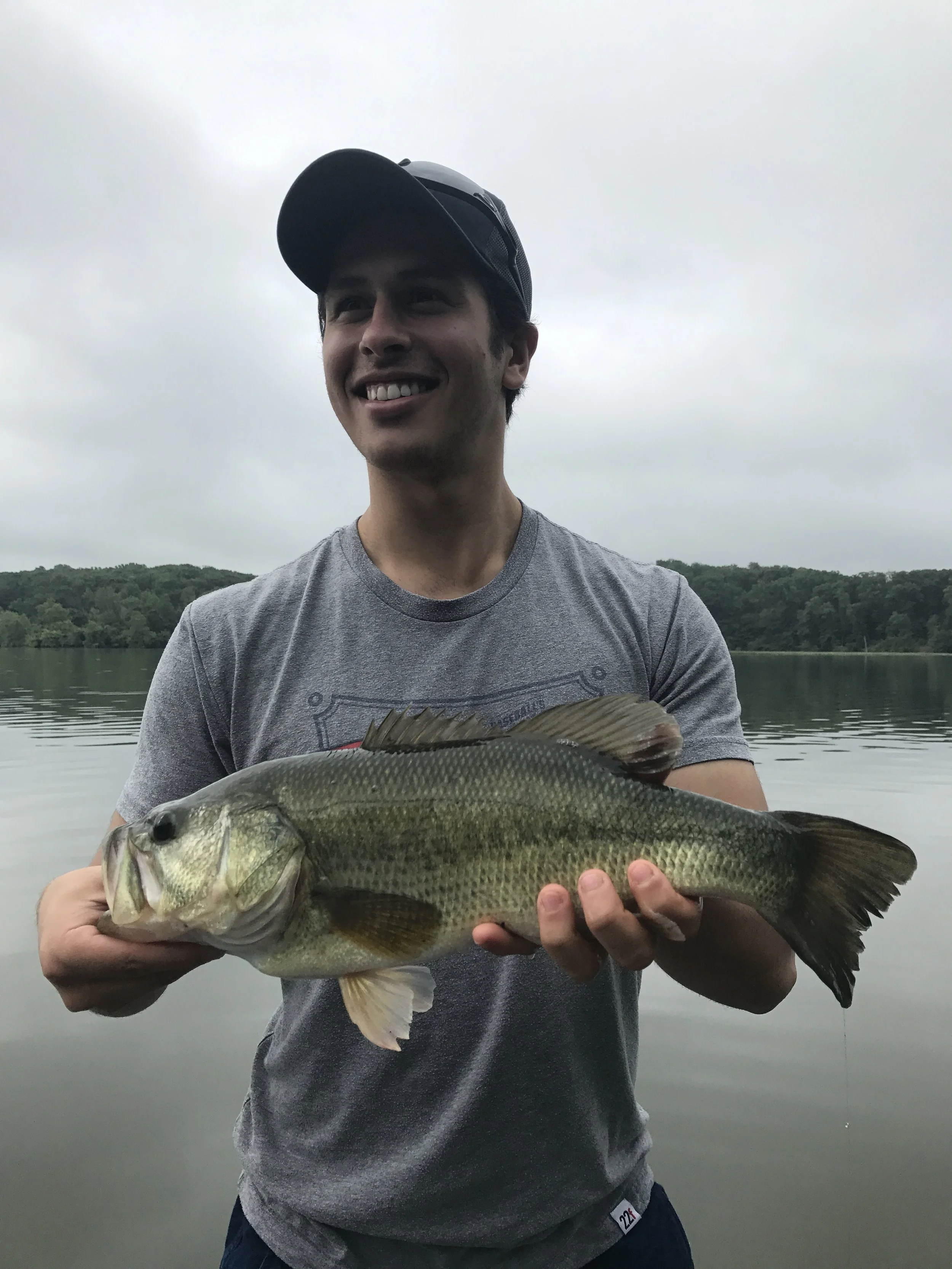 Young man wearing a cap and gray t-shirt smiling while holding a large fish in front of a lake with trees in the background on a cloudy day.