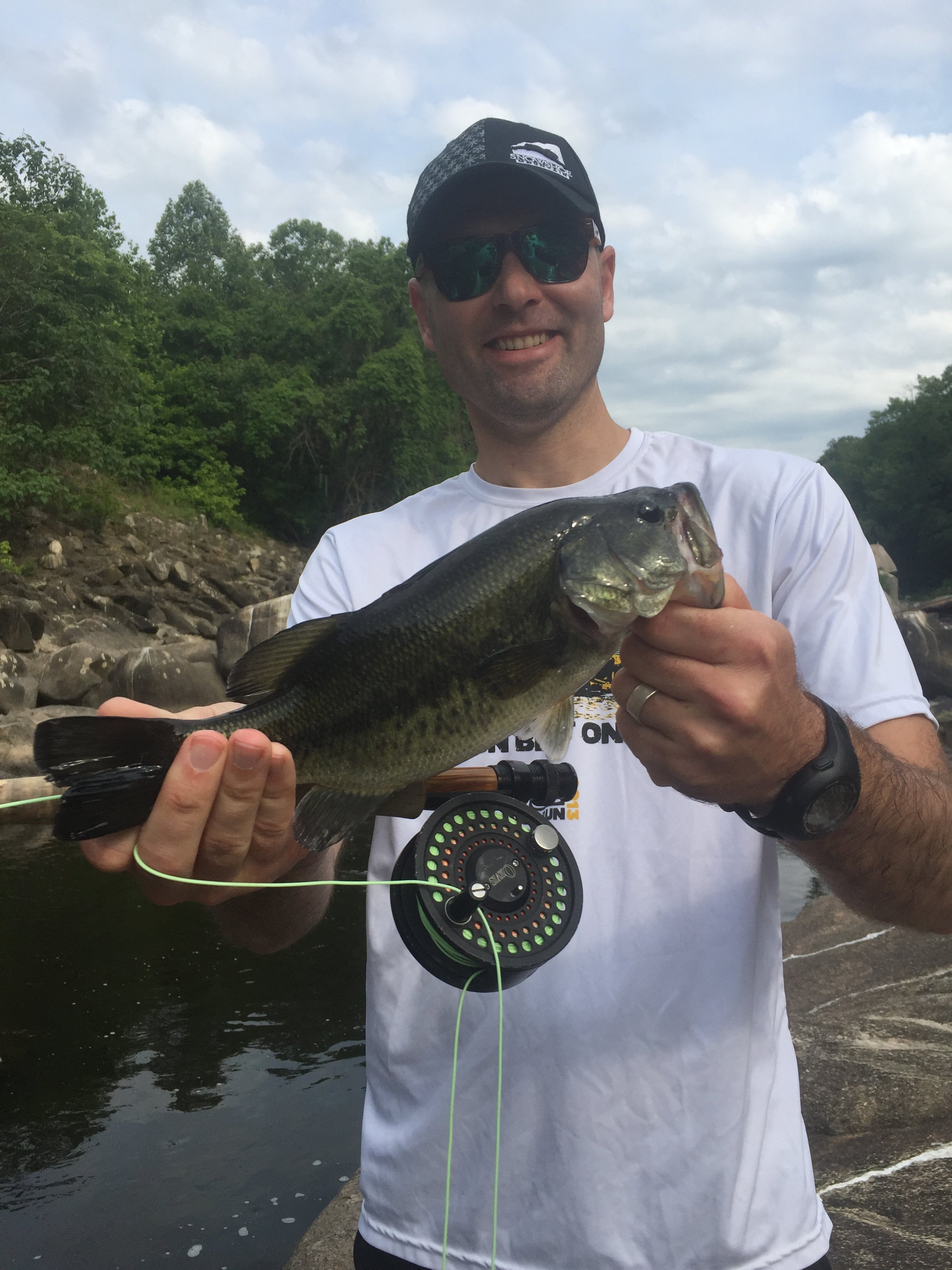 Man in sunglasses and a white T-shirt holding a large fish and smiling outdoors near a rocky riverbank with trees and cloudy sky in the background.