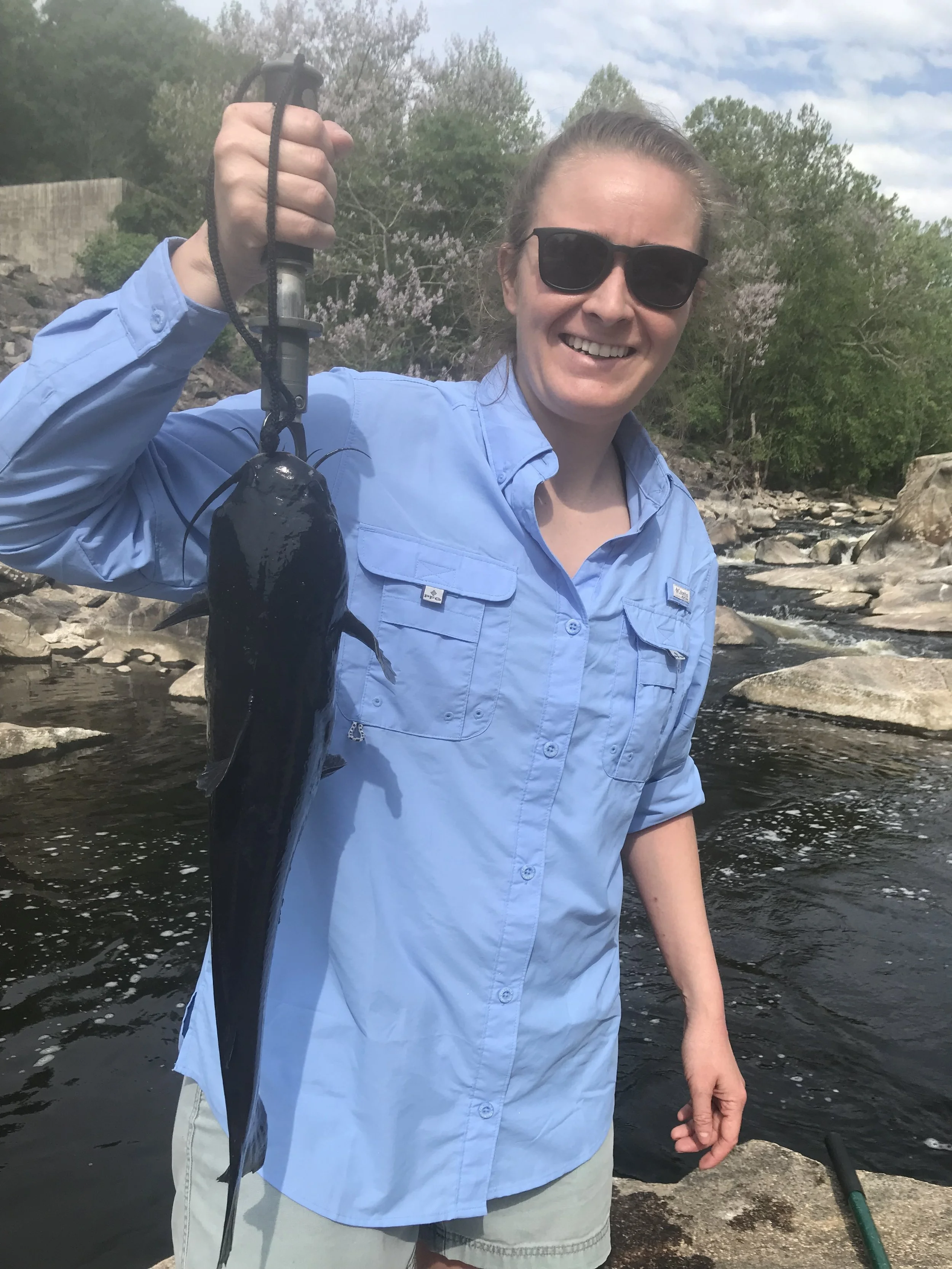 Woman with sunglasses smiling, holding a fish she caught, standing by a river with rocks and greenery in the background.