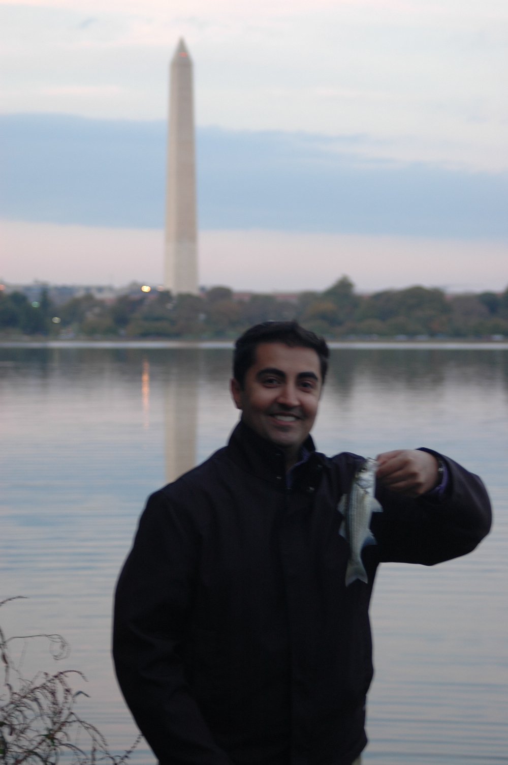 Man holding a fish by a body of water with a tall monument in the background