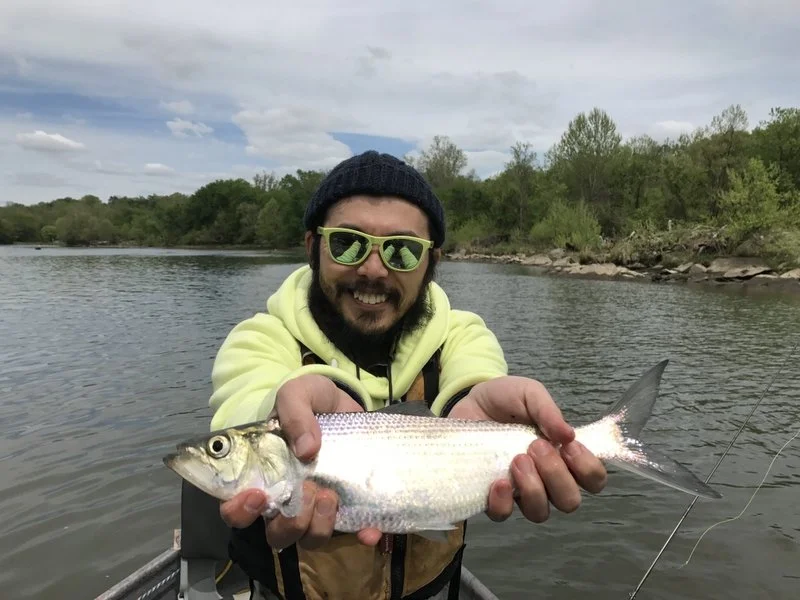 Man smiling and holding a fish over a river with trees in the background.
