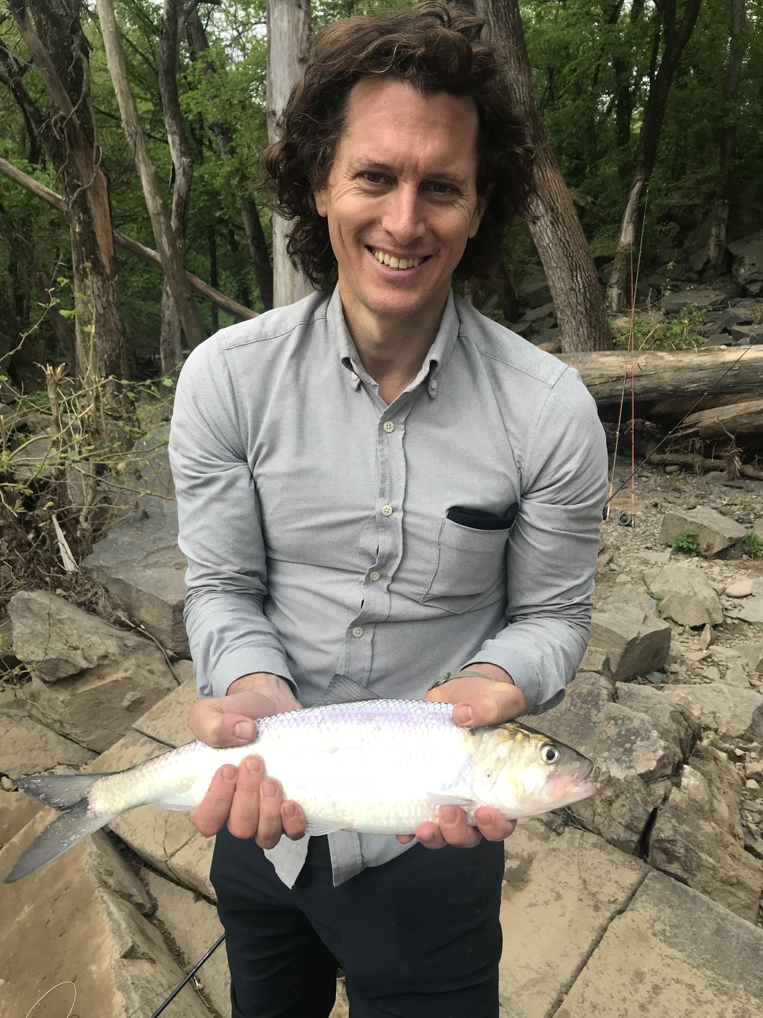 A man with curly brown hair smiling and holding a fish in an outdoor setting with trees and rocks.