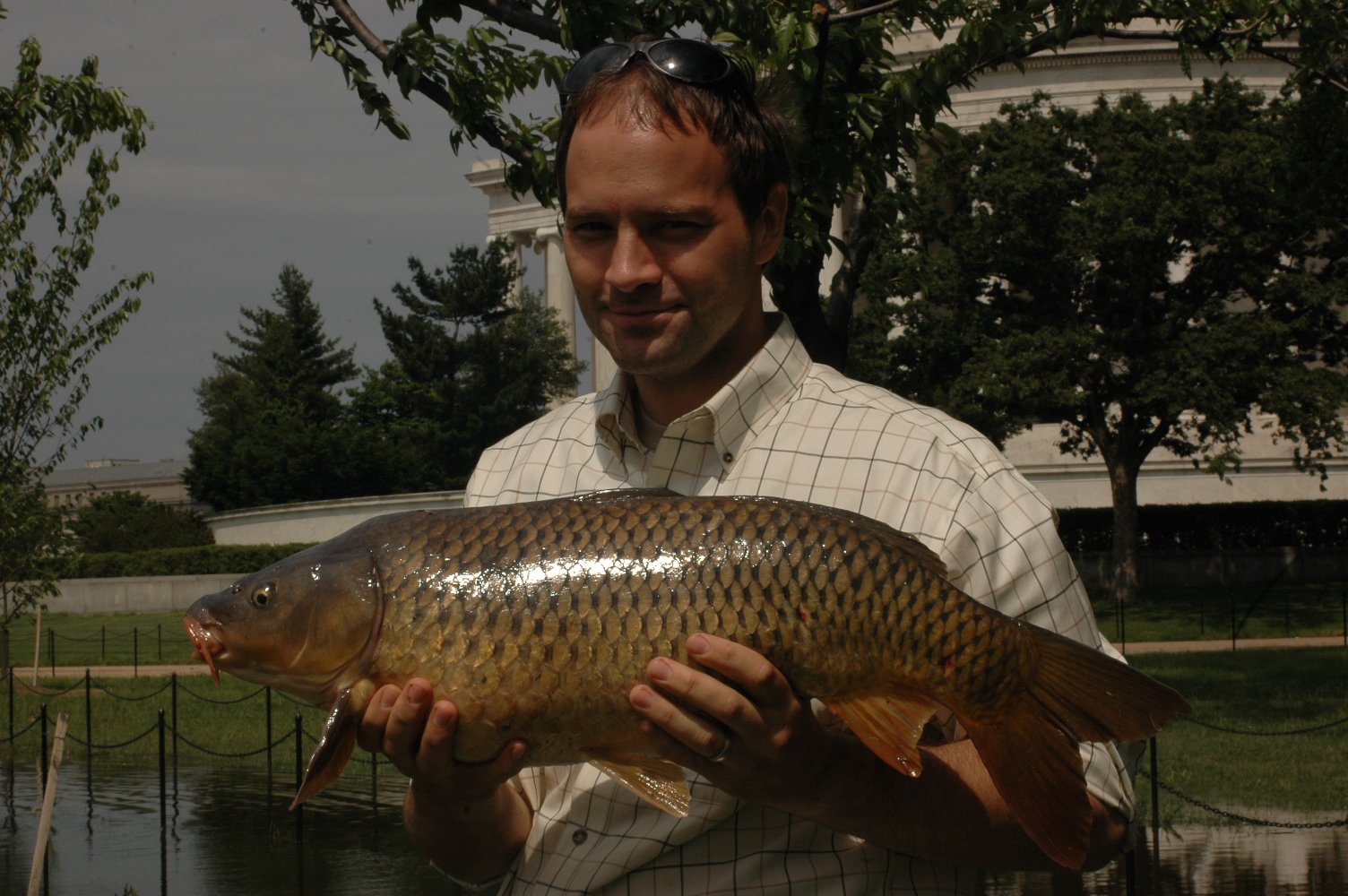 Man holding a large fish outdoors near water, with trees and a white building in the background.