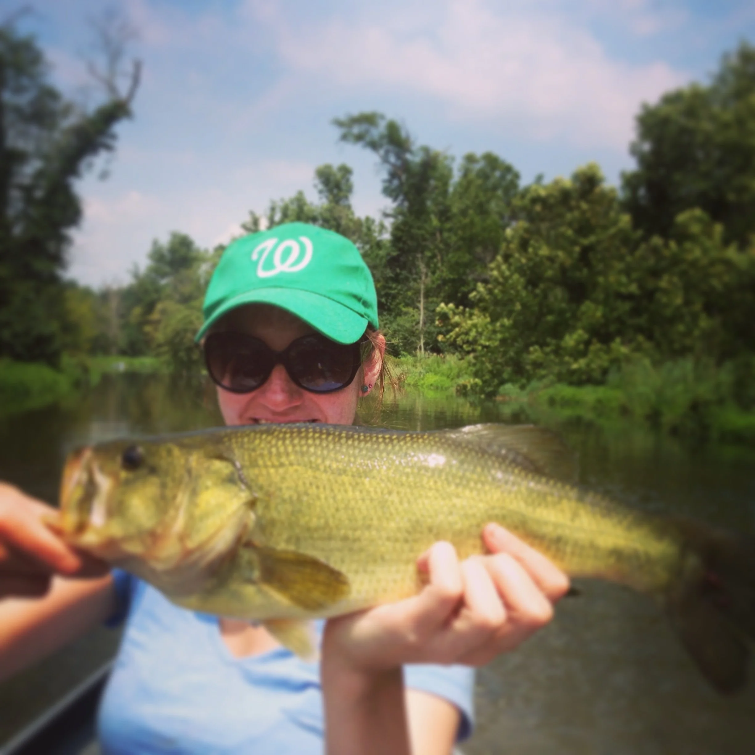 Woman wearing sunglasses and a green baseball cap holding a large fish with greenish-yellow scales near a river and lush trees in the background.