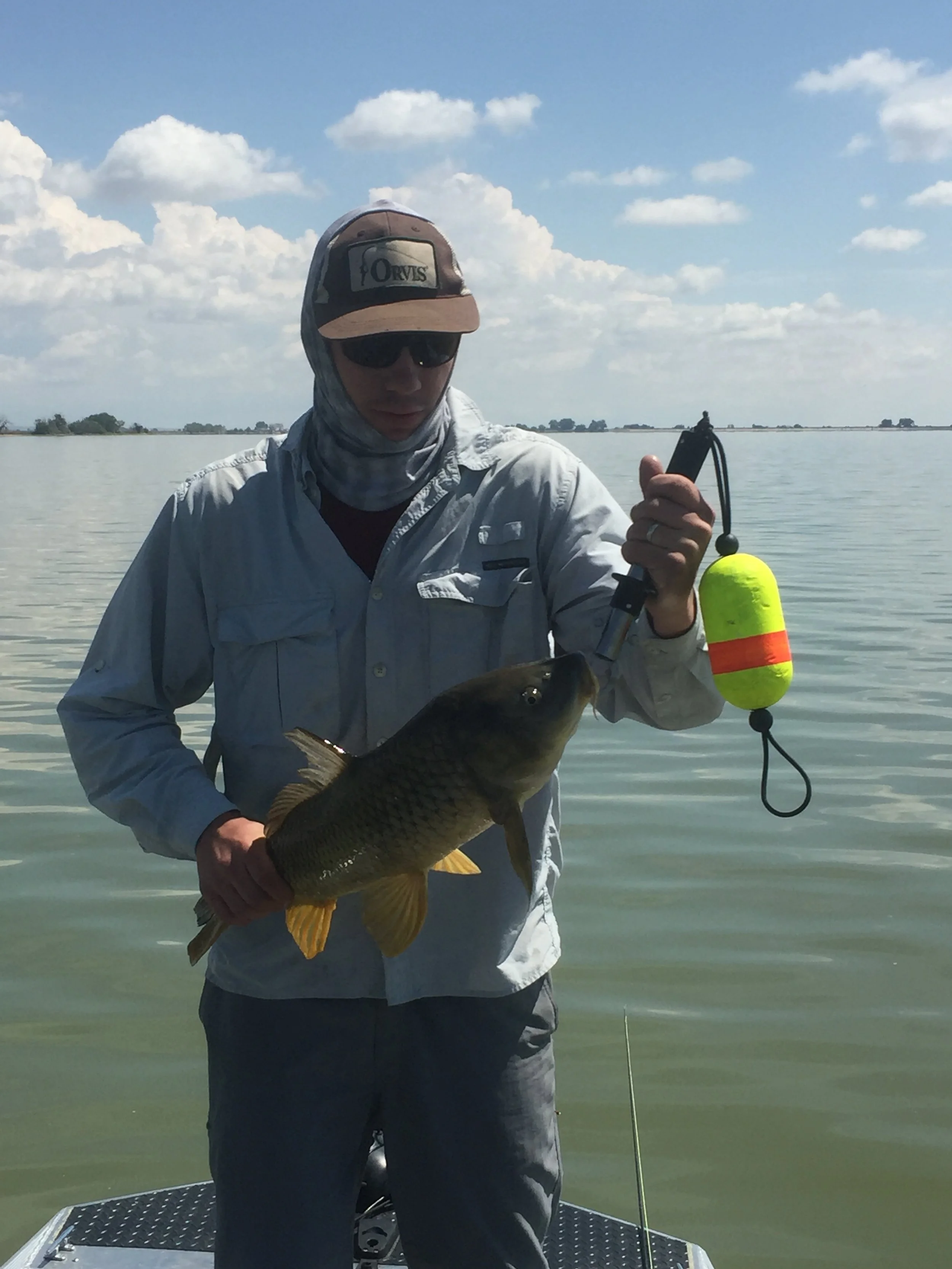 A person wearing a hat, sunglasses, and a long-sleeve shirt holding a large fish on a boat in a body of water under a partly cloudy sky.