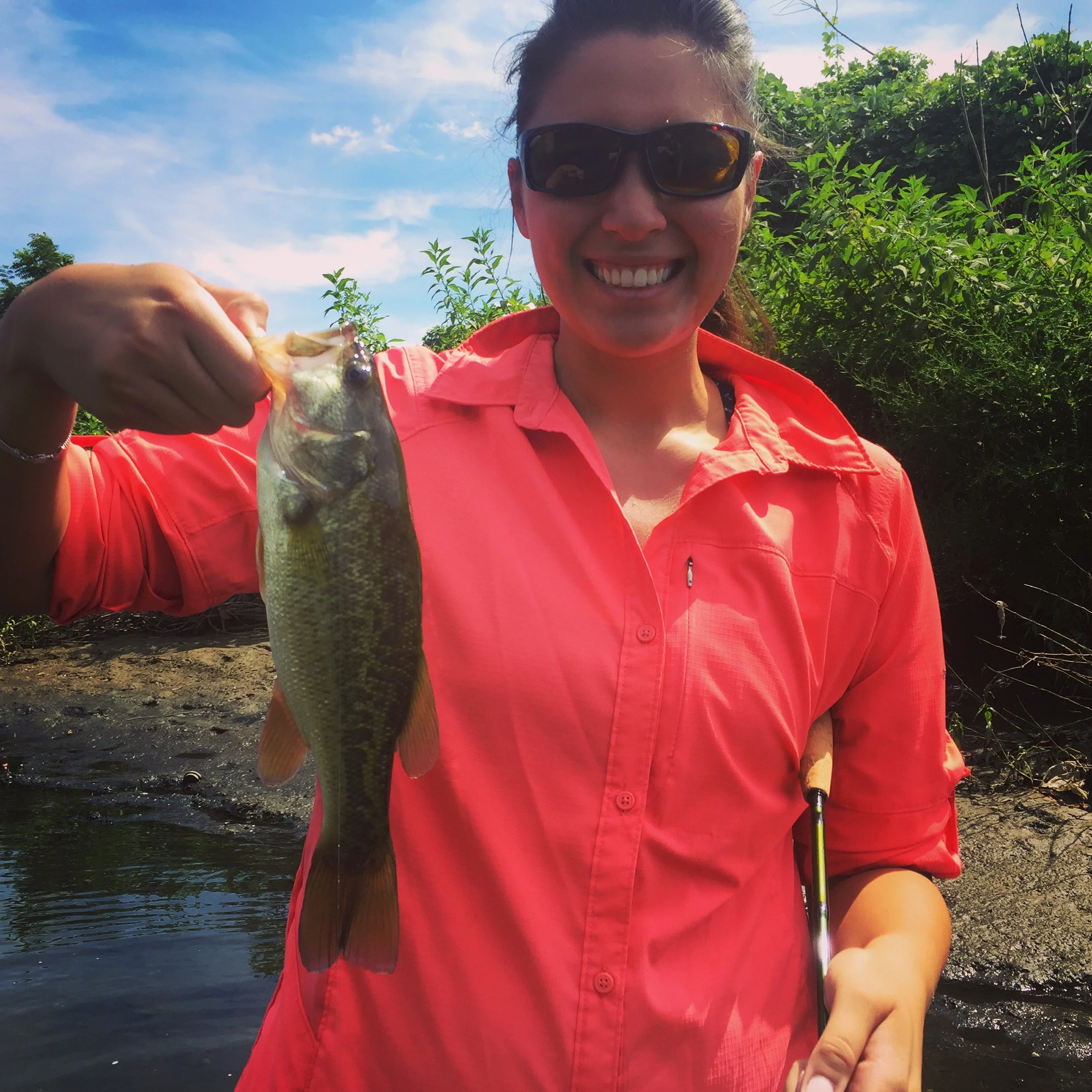 A woman in a bright coral shirt and sunglasses standing outdoors near water, holding a fish she caught, smiling.
