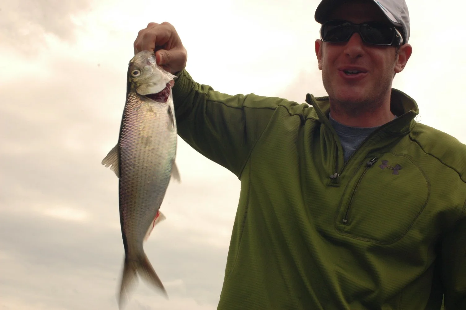 Man wearing sunglasses and a green jacket holding a fish with grey and pinkish scales against cloudy sky background.