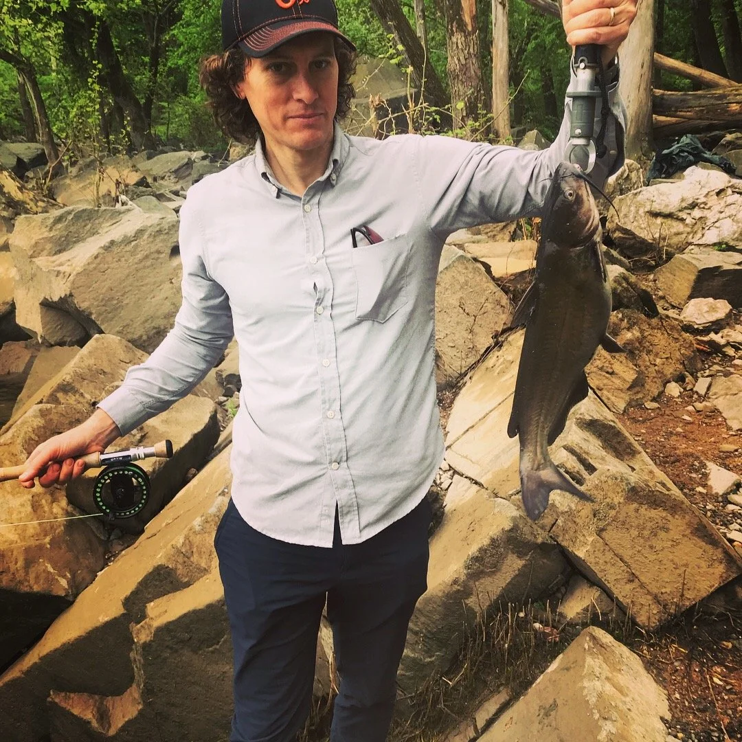 A man holding a large fish with a fishing rod in an outdoor rocky area.