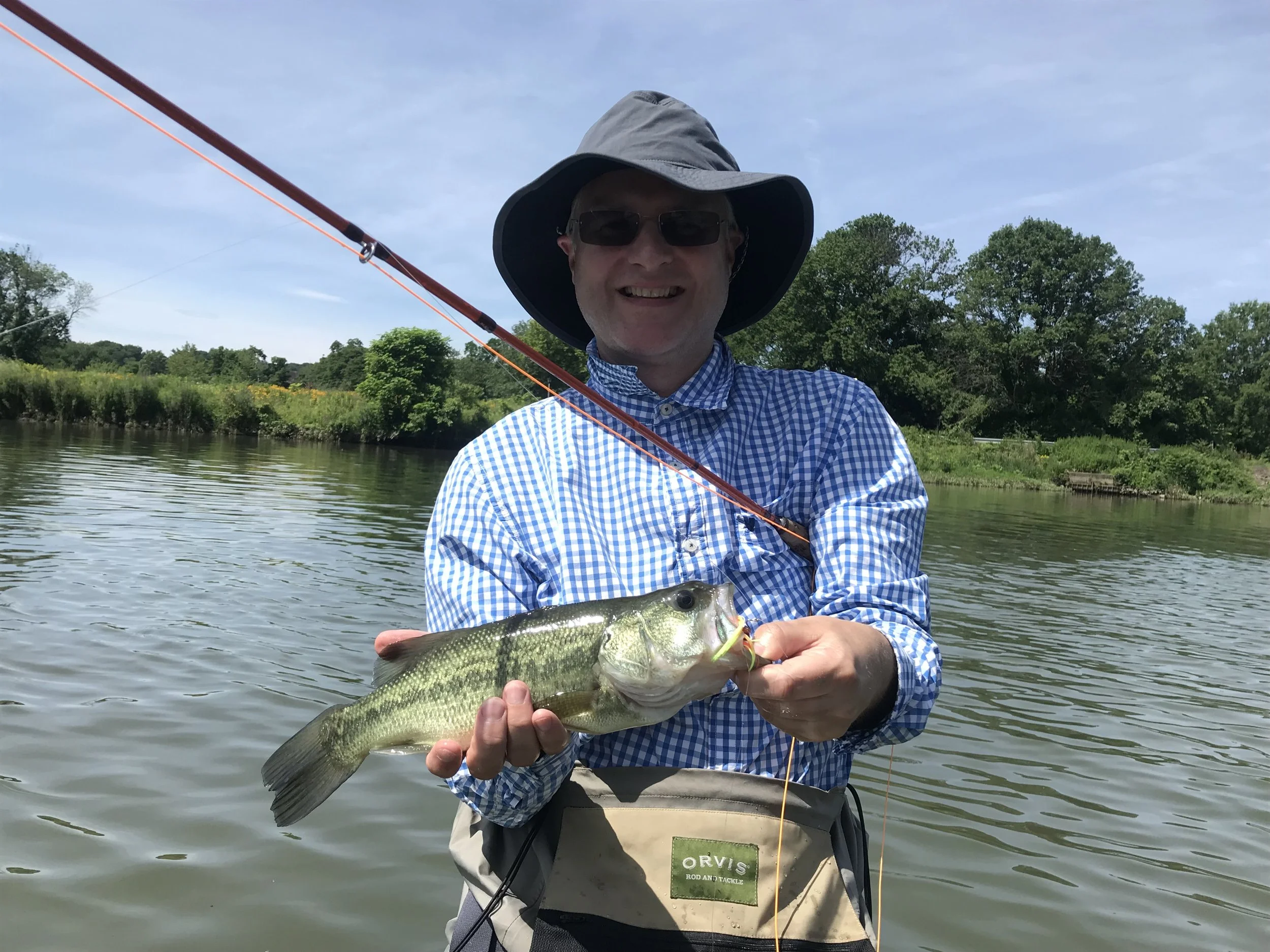 Man in hat and sunglasses holding a largemouth bass fish he caught while fishing on a river on a sunny day with green trees in the background.