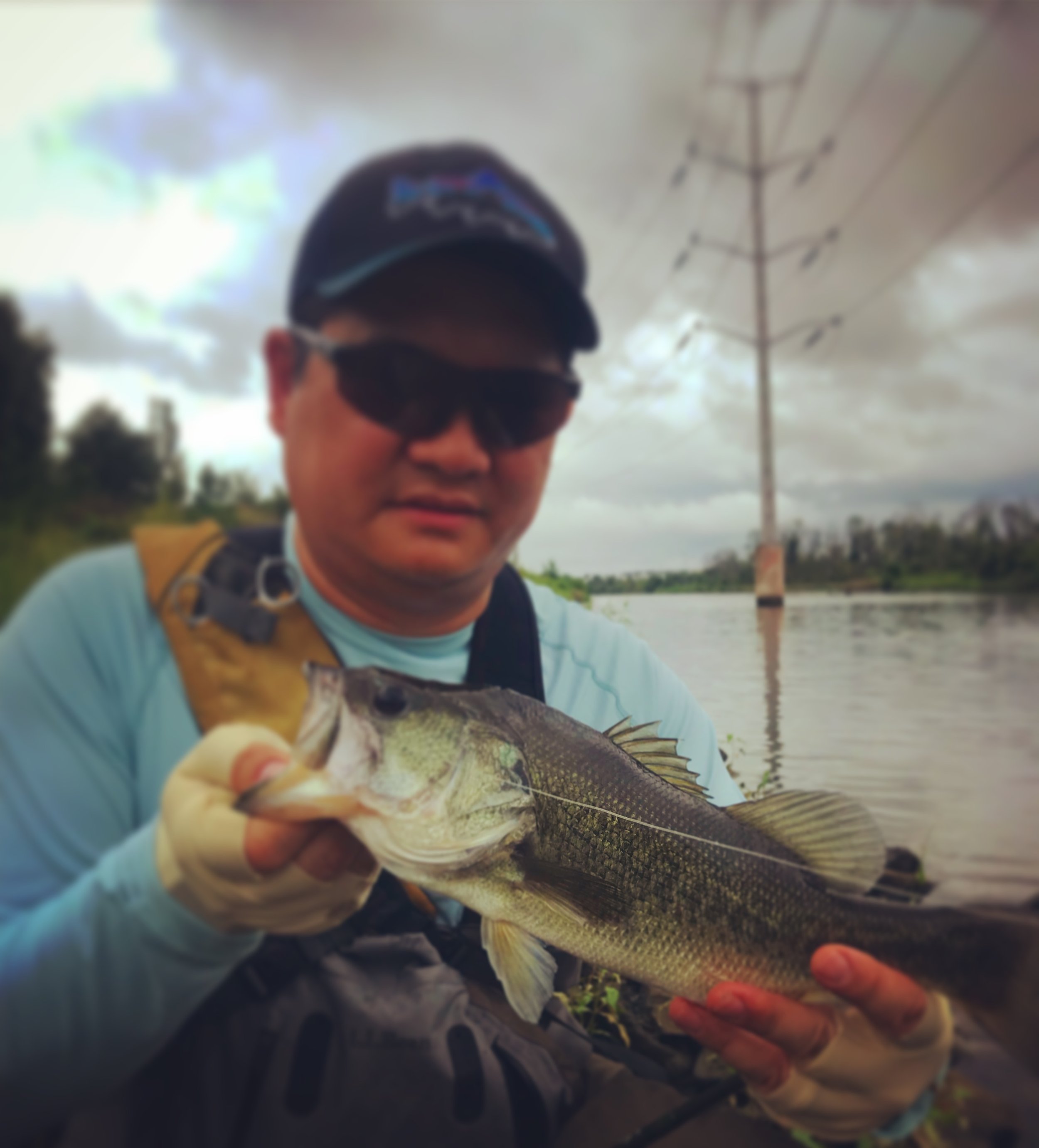 Man wearing sunglasses holding a largemouth bass fish near a river.