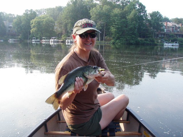 Woman in a boat holding a fish she caught, smiling, with water and trees in the background.
