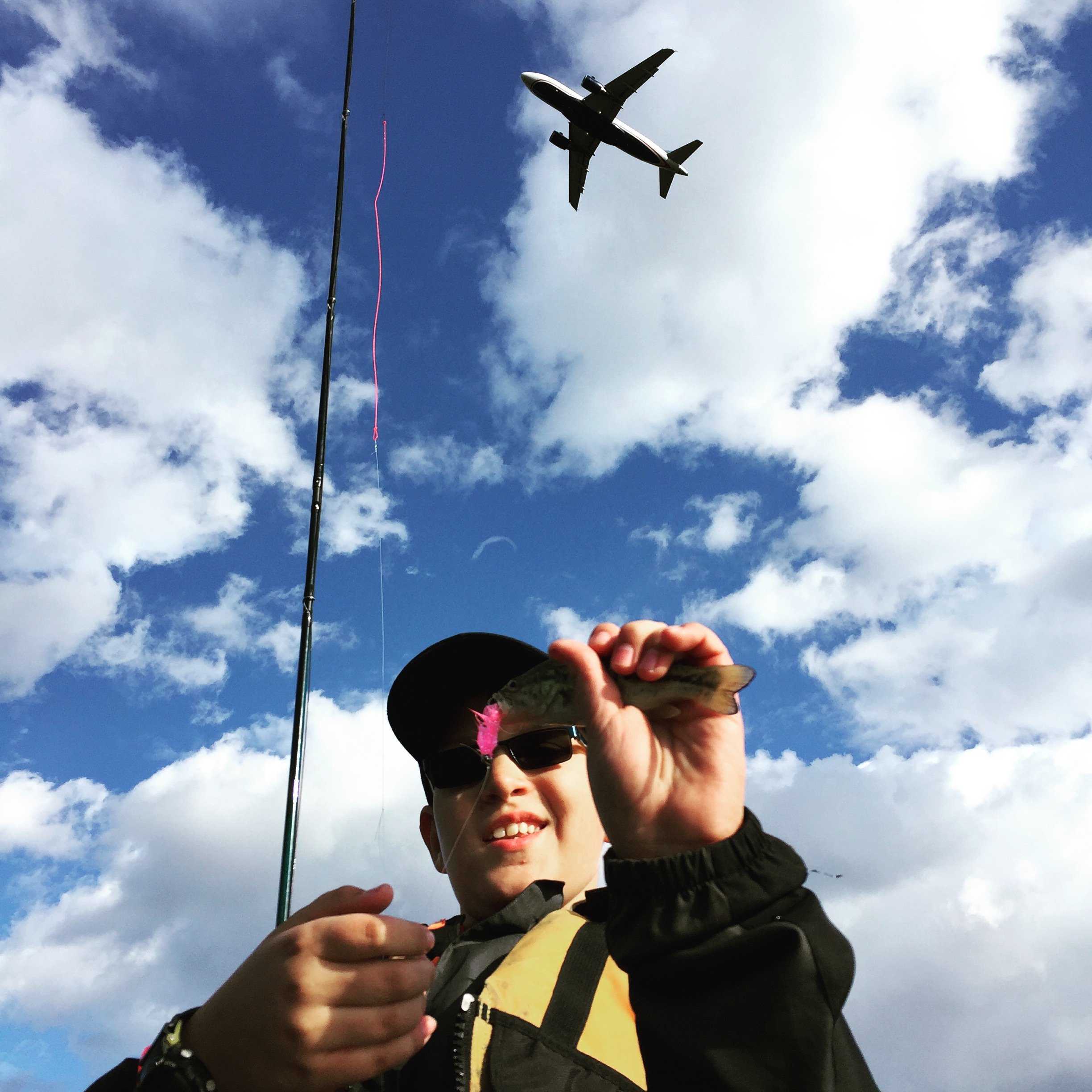 A boy wearing a cap, sunglasses, and a fishing vest is holding a small fish with a pink tag in front of a blue sky with scattered clouds. An airplane is flying overhead and a fishing pole is visible.