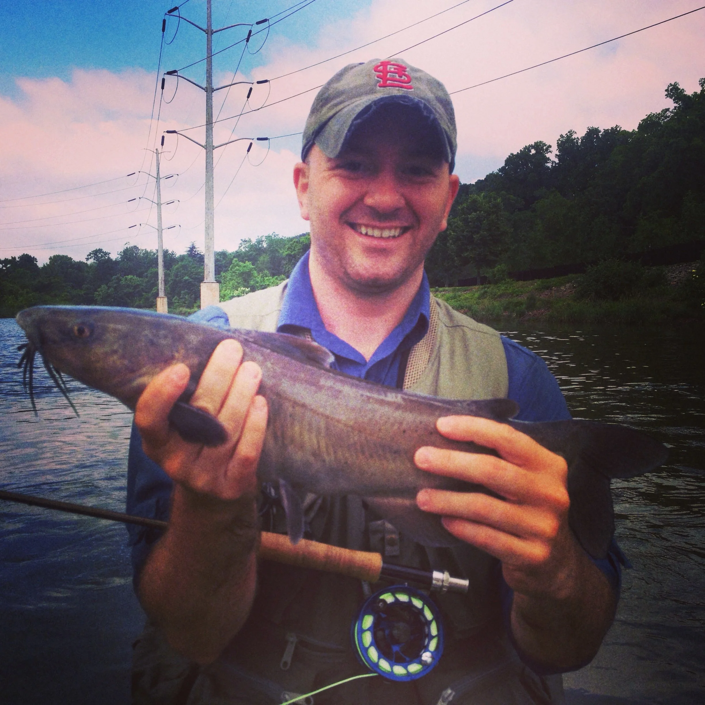 A smiling man wearing a St. Louis Cardinals cap holding a fish near a river with power lines and trees in the background.