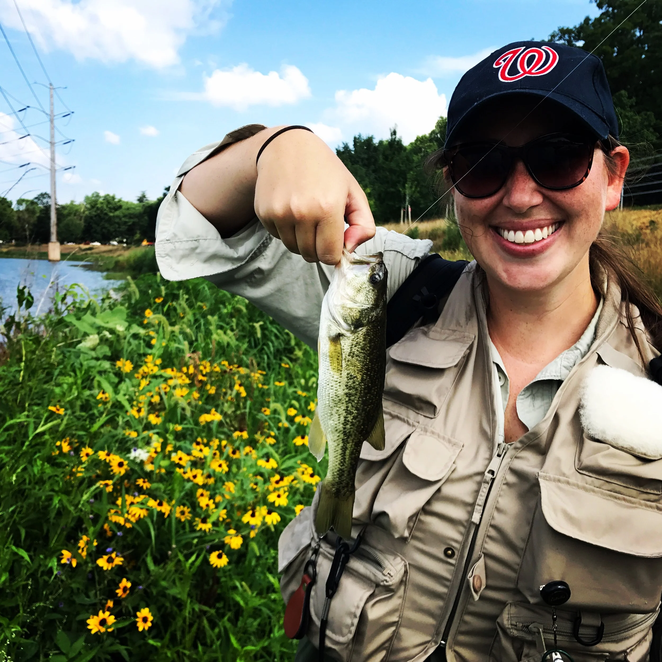 Woman holding a fish near a pond with yellow flowers and green vegetation, wearing a beige fishing vest and cap with a red 'W' logo, sunglasses, smiling outdoors on a sunny day.