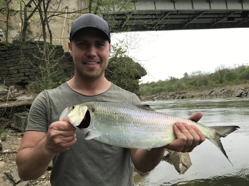 A young man wearing a baseball cap and a gray T-shirt holding a large fish he caught by a river, with a bridge overhead in the background.