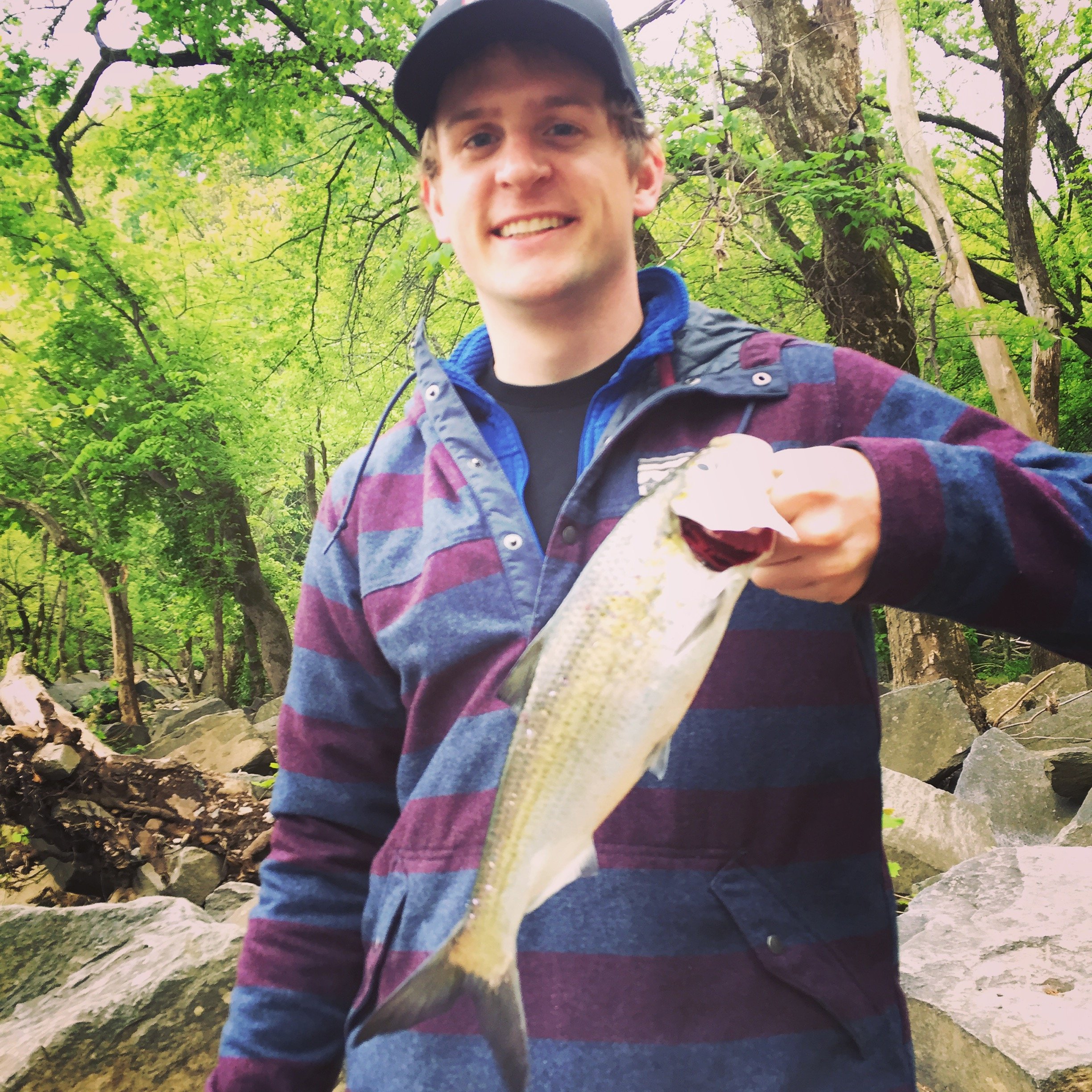 A young man smiling while holding a fish he caught in a river surrounded by green trees and rocks.