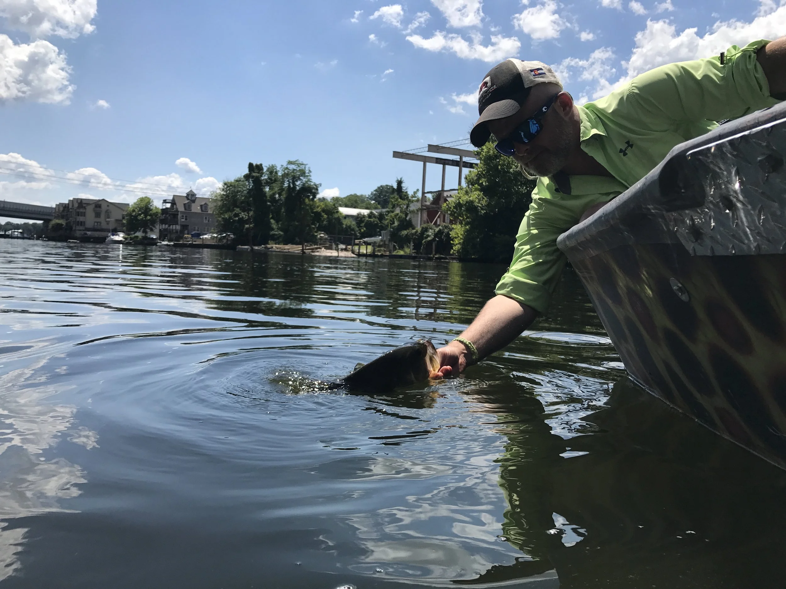 Man in a neon green shirt and sunglasses extends his arm from a boat to hand a fish back into the water on a sunny day with blue skies and scattered clouds, on a river with houses and trees in the background.
