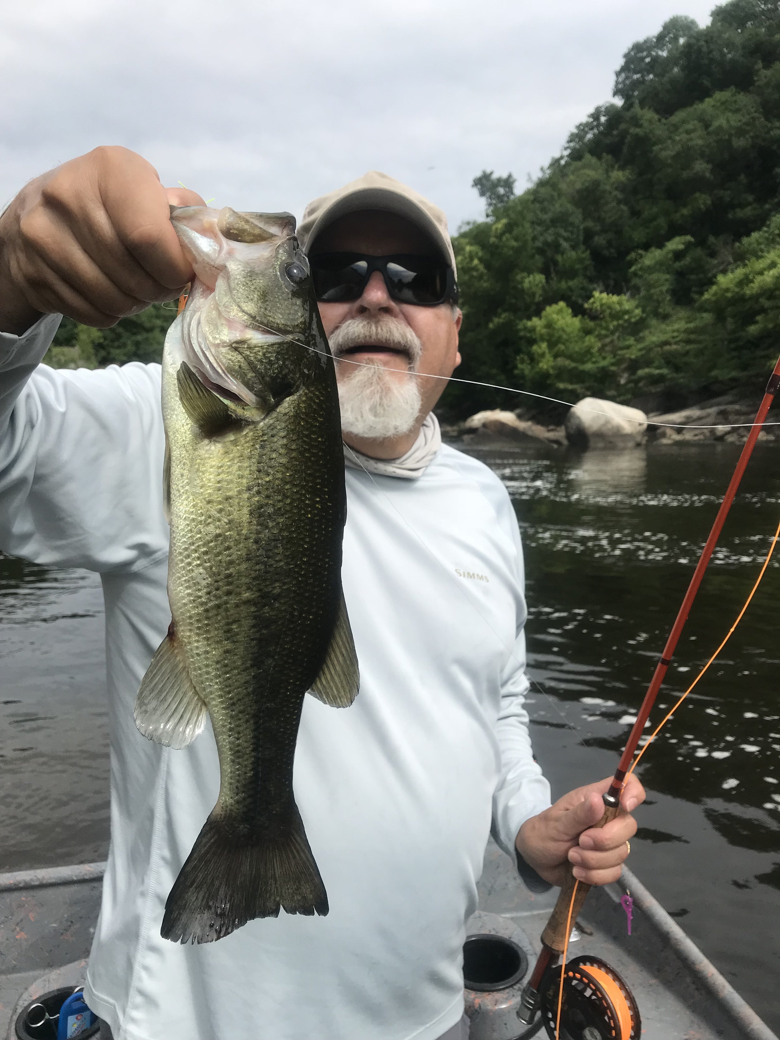 Man in outdoor clothing holding a large fish he caught while fishing on a river with trees and rocks in the background.