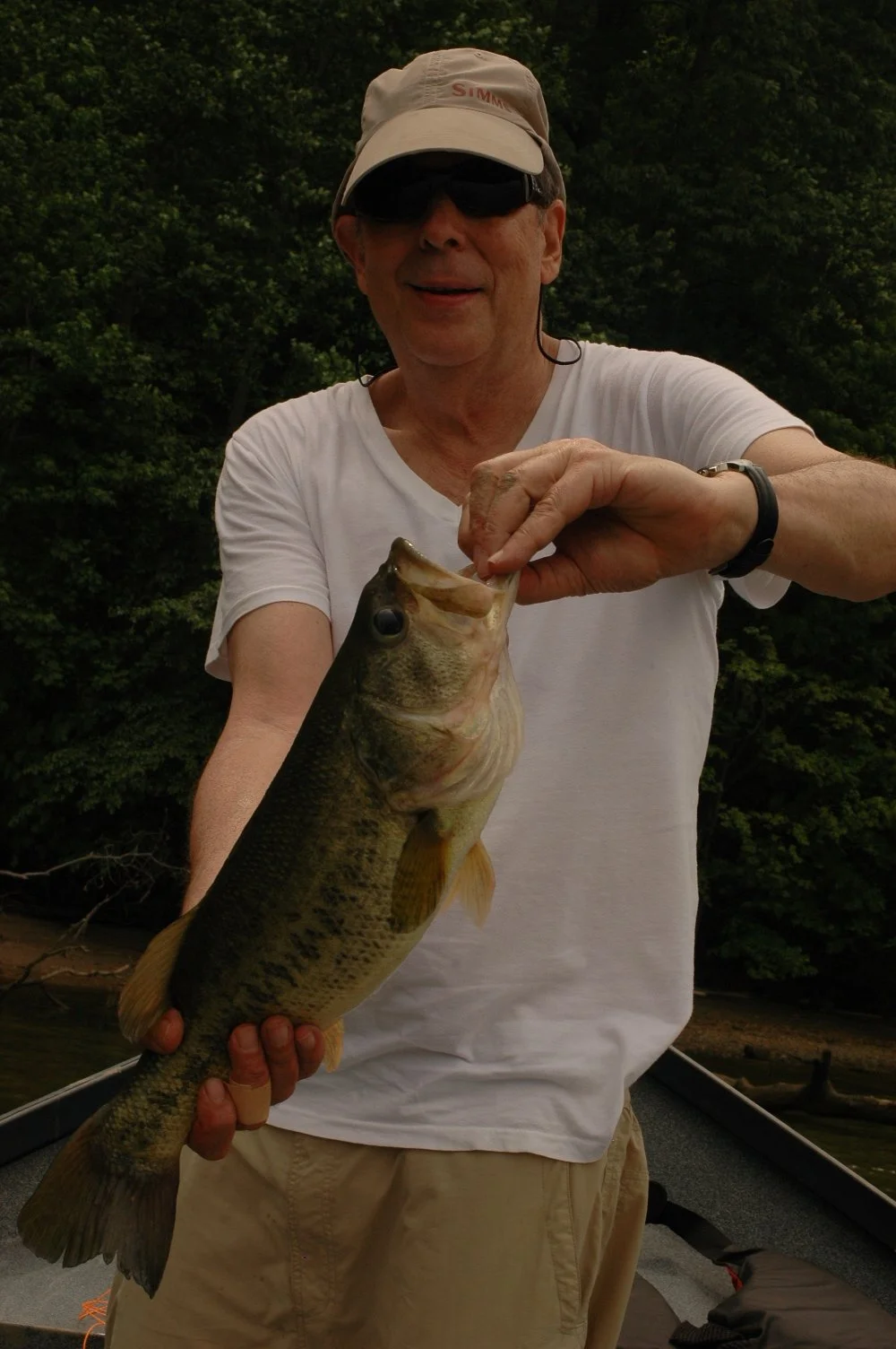 A man wearing a tan cap, sunglasses, and a white t-shirt is holding a largemouth bass fish with one hand, showing it to the camera. He is outdoors with a background of green trees.