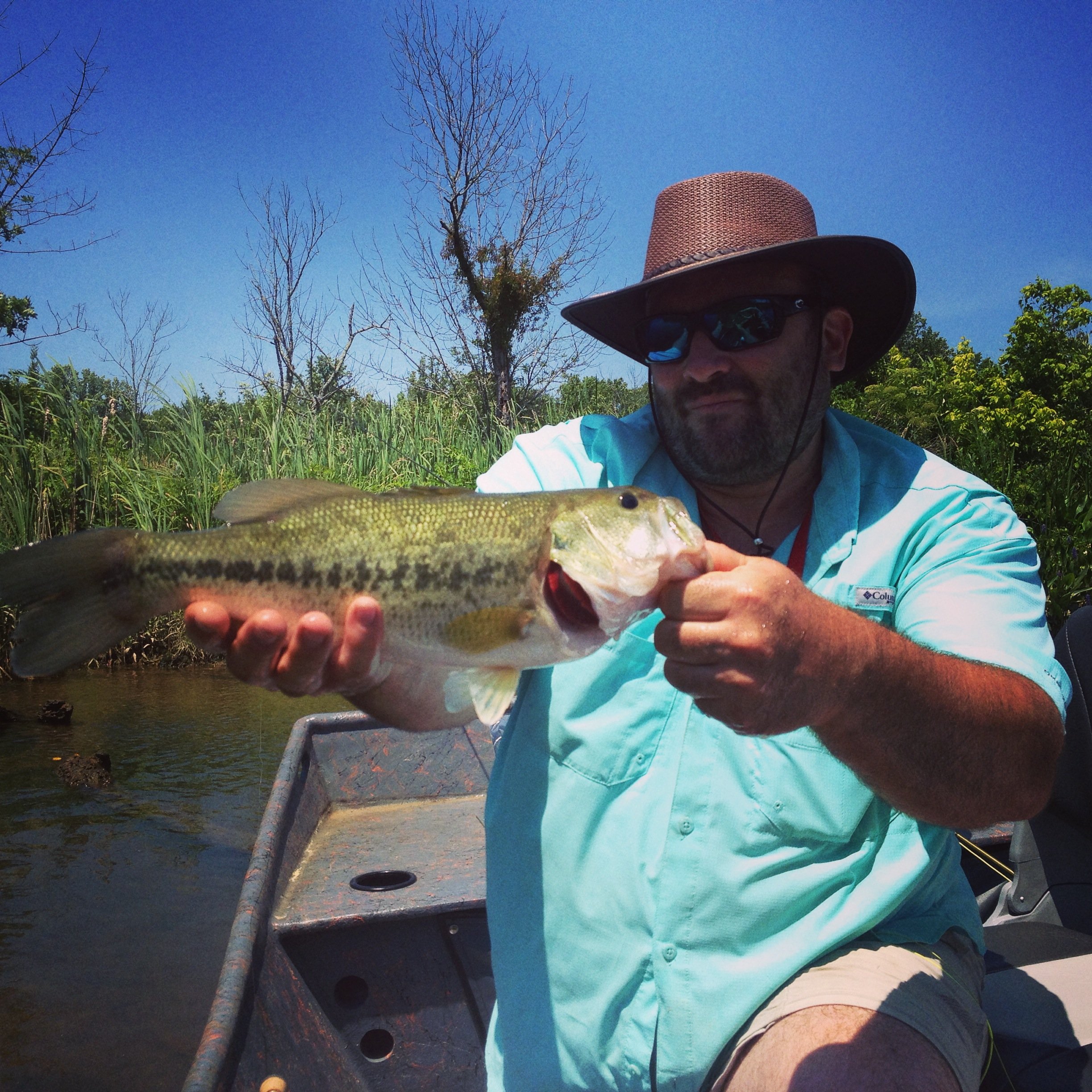 Man wearing sunglasses and a wide-brimmed hat holding a largemouth bass fish on a boat near a river with trees and blue sky in the background.