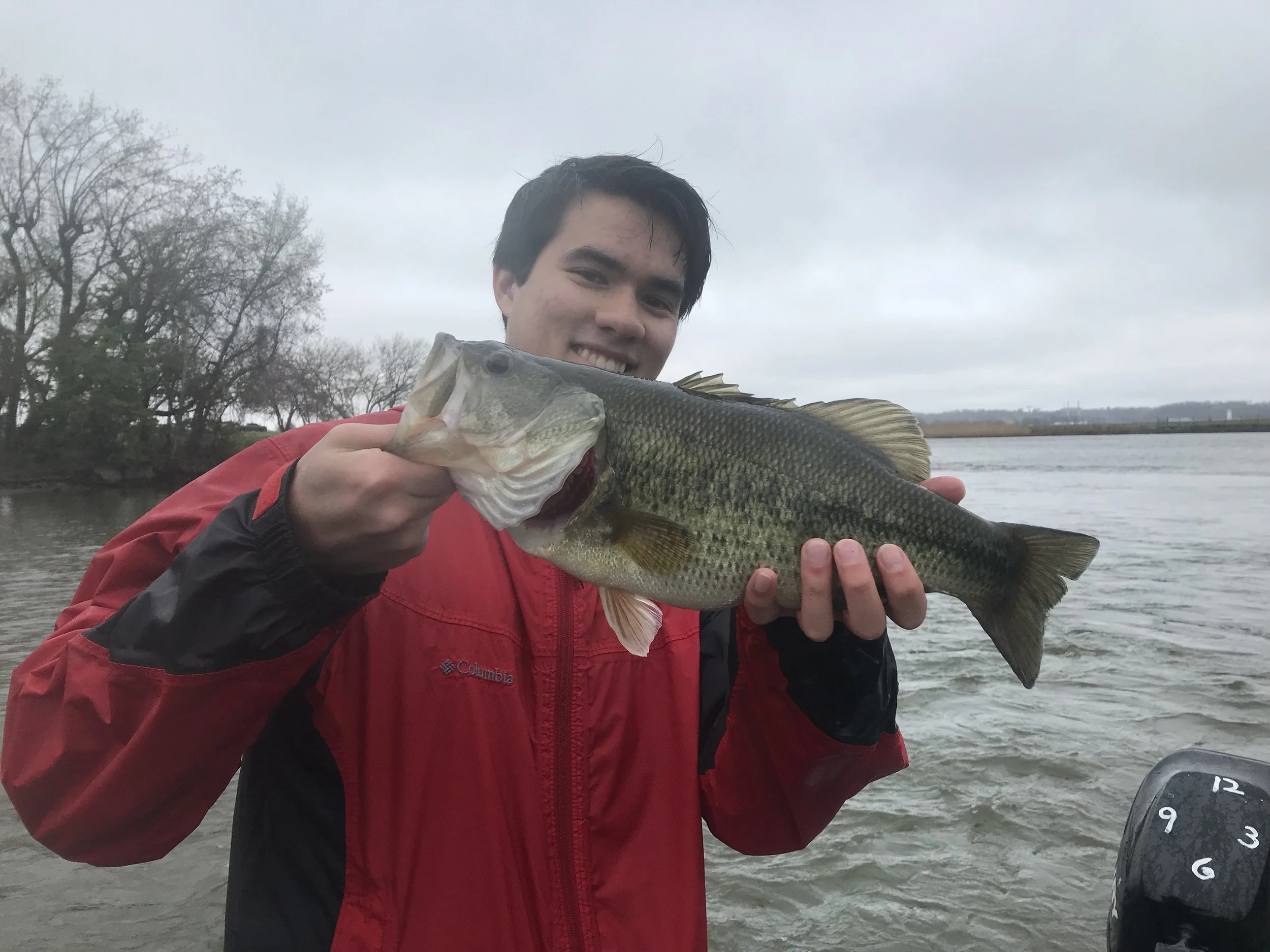 A man in a red jacket holding a large freshwater fish, possibly a bass, near a body of water on a cloudy day.