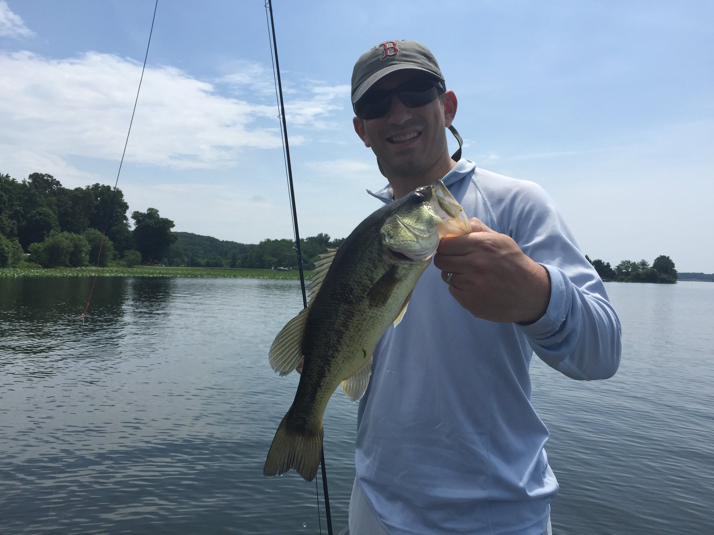 Man wearing sunglasses, a cap, and a light-colored shirt holding a largemouth bass fish on a fishing line near a lake with trees and hills in the background.