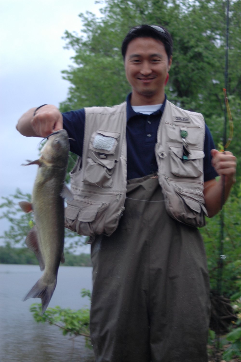 Man smiling while holding a fish he caught, standing outdoors near a river and trees.