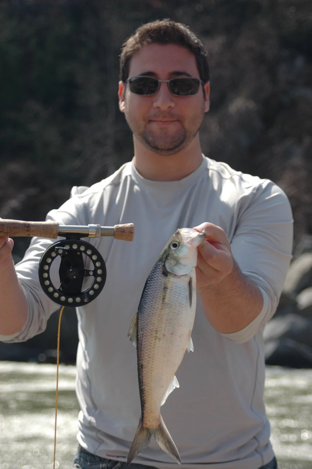 Man wearing sunglasses and a white shirt holding a fishing rod and a large fish outdoors near a river.